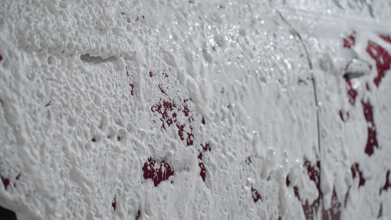 Detailed close up of thick soapy foam splashing and spreading across red car surface during professional washing process showing texture of dense bubbles forming white layer over vehicle exterior