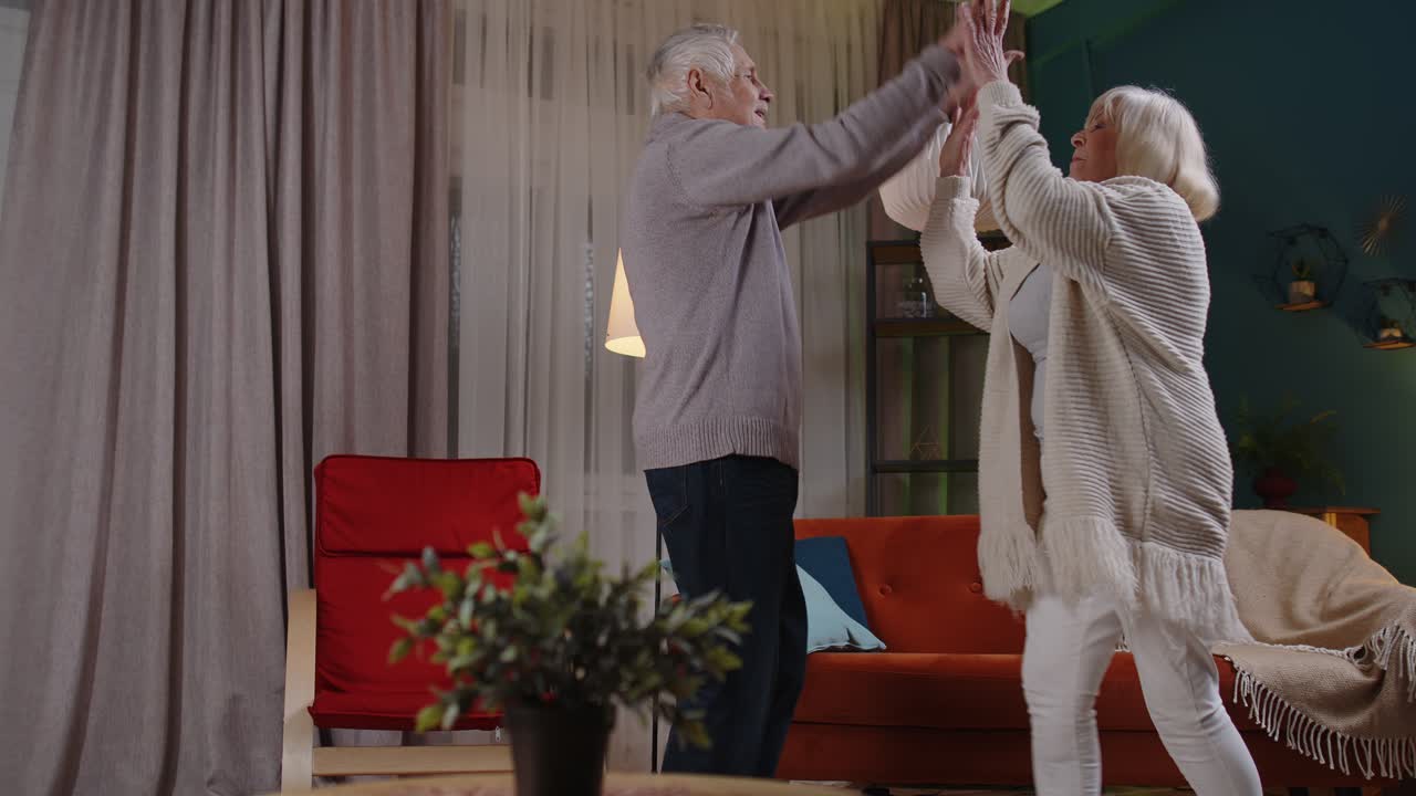 걱정 없는 노인 커플이 집에서 춤을 추고 휴식을 취하고 현대 음악으로 재미를 느끼고 집에서 웃고 있습니다.