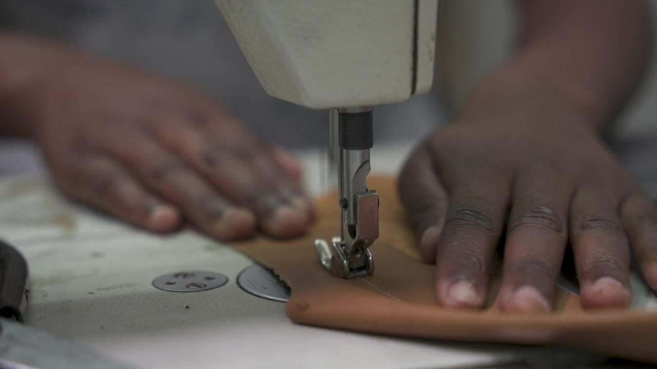 Close-up of hands maneuvering leather beneath a sewing machine needle in a workshop. Captures the precision and craftsmanship of leatherworking. Suitable for artisan or craft-focused projects.