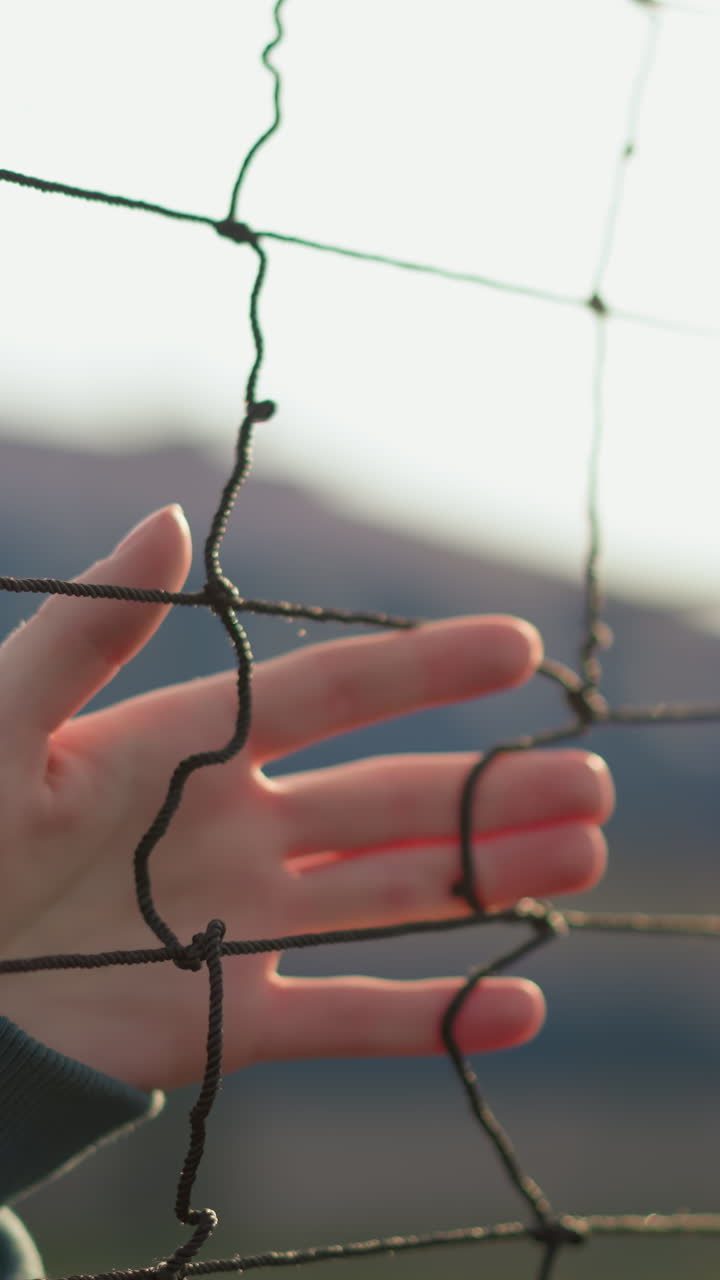 초록색 스웨터를 입은 여자가 배구망 위에 손을 미끄러뜨리고 빛이 그녀의 얼굴에서 반사되며, 손의 움직임에 초점을 맞추고, 부드러운 낮빛으로 배구장의 흐릿한 배경