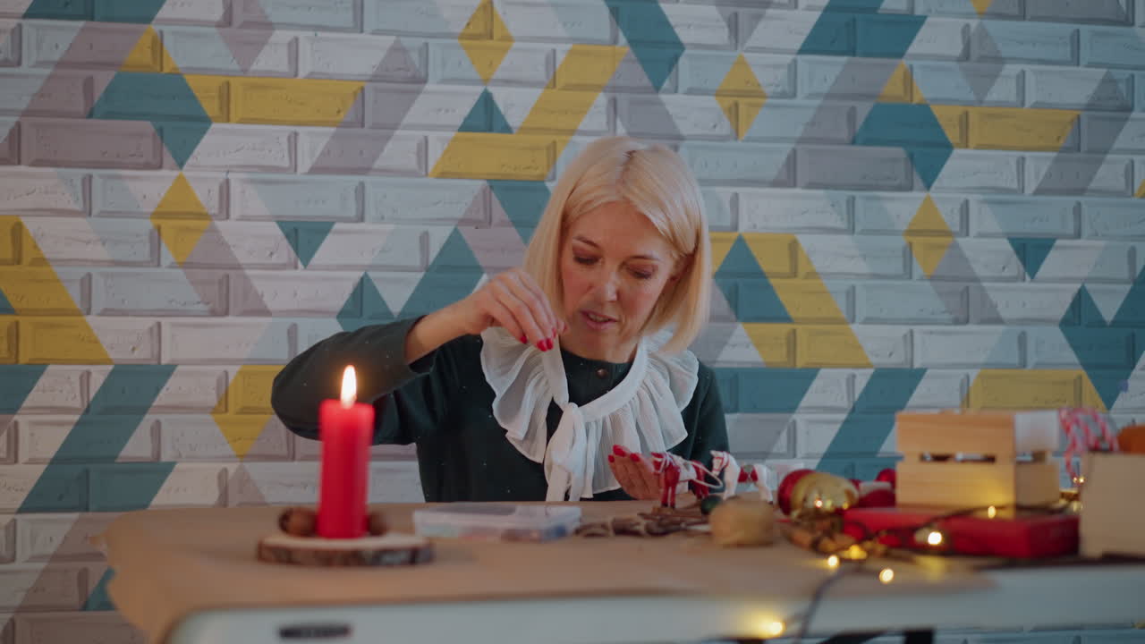 Donna caucasica che incarta un regalo di Natale a lume di candela su un tavolo accogliente, con nastro, forbici, piccole scatole, lucine fatate e una parete a motivi. L'anziana sorride concentrata mentre crea una decorazione festiva, avvolta da un caldo bagliore.