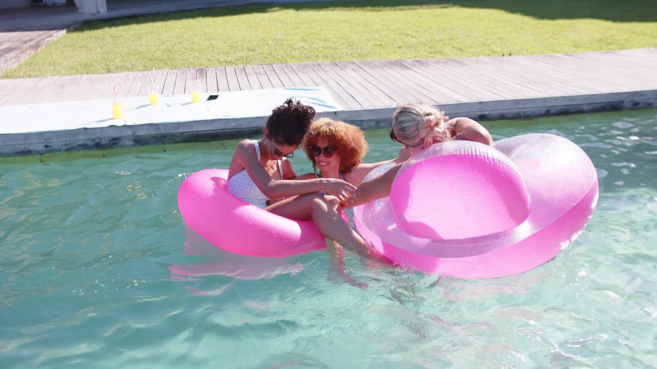 Three women in swimwear laughing, third woman reaching and pulling pink floats connecting in pool