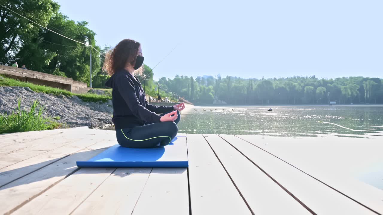 Young curly woman practicing yoga near the lake while wearing black protective mask. Sunny day. Meditating in lotus position on the yoga mat. Corona Virus idea. Chisinau, Moldova