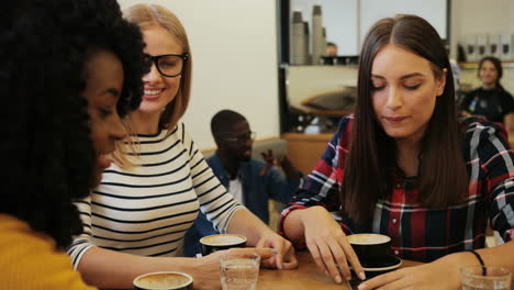 close-up view of african american and caucasian women friends laughing and watching something on a smartphone sitting at a table in a cafe