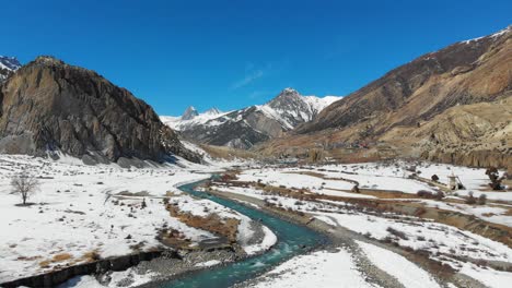 wide drone shot of a river surrounded by snow on a bright sunny day in manang, nepal.