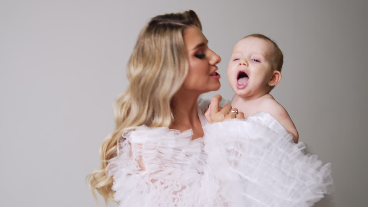 Beautiful woman in white robe holding a lovely naked child. Mother touches baby's nose and cheeks. White backdrop.