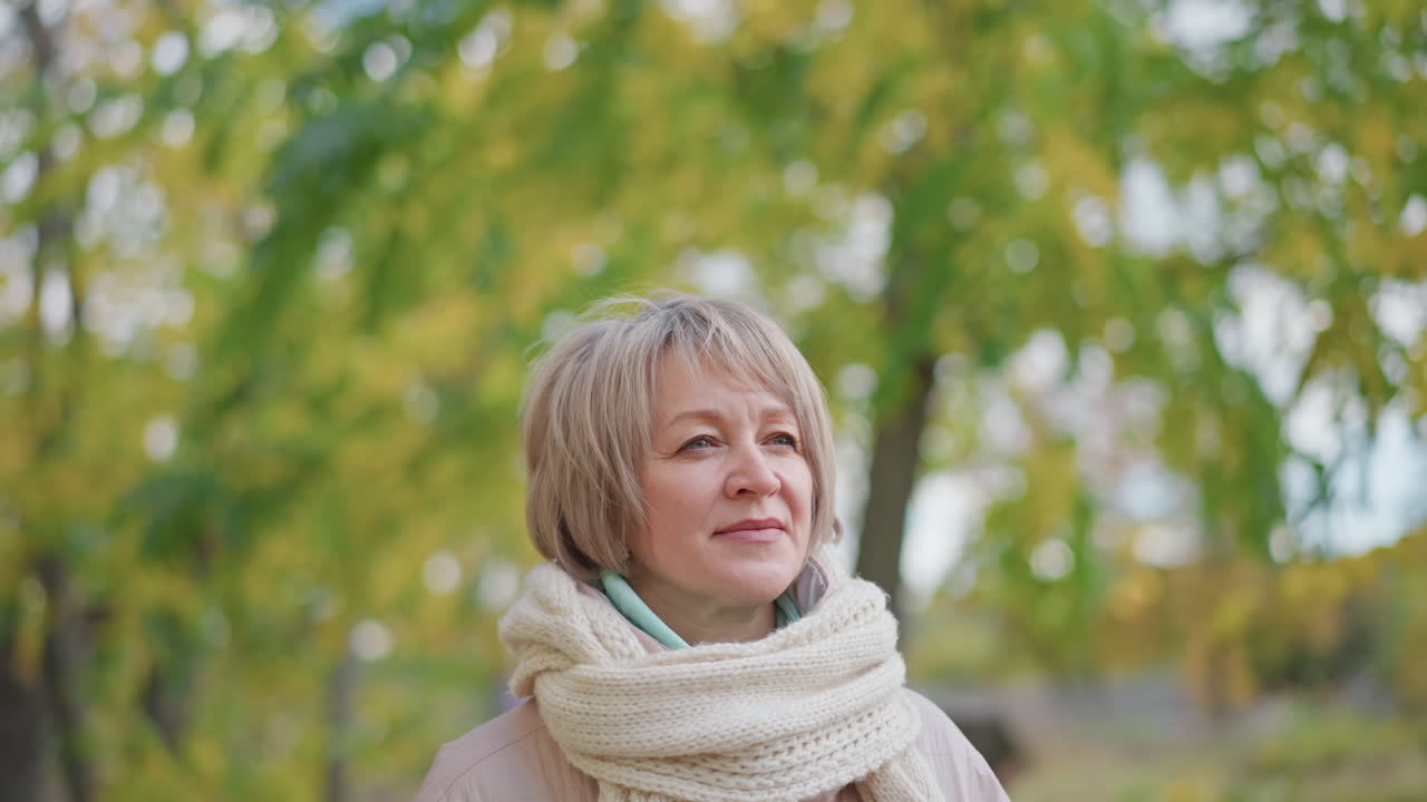 elegant mature woman wrapped in thick scarf stands outdoors with eyes closed, appearing calm and thoughtful while surrounded by blurred golden green foliage on peaceful autumn day