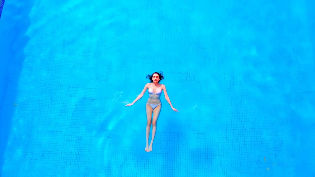 Top-down motion of woman floating supine in tiled turquoise pool, moving slowly with gentle limb adjustments, soft ripples, and relaxed body posture under bright sunlight, creating calm wellness mood