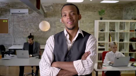 young african businessman is standing with arms crossed and watching at camera in office, smiling, colleagues are working with laptops, work concept