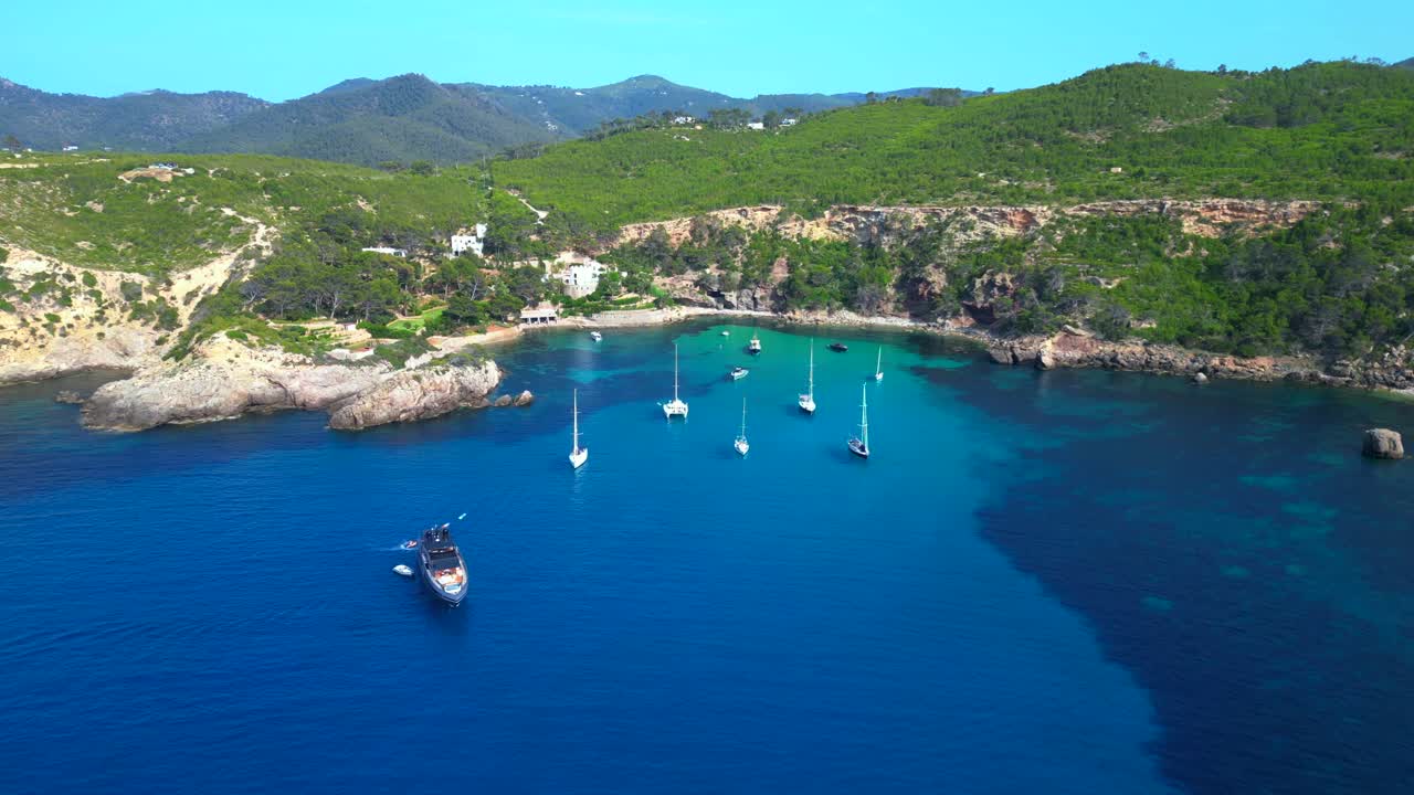 sailboats and yachts moored in turquoise waters of a bay on Ibiza, Spain, surrounded by lush greenery. Wonderful aerial view flight drone shot footage from above