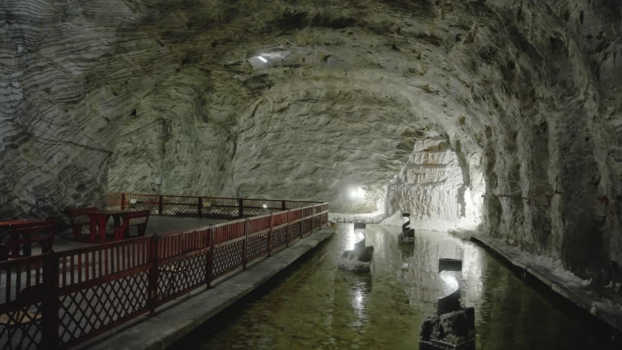 A static shot inside the massive Targu Ocna salt mine. A large cavern features rough-hewn salt walls, a wooden railing, and a pool of water reflecting the vaulted ceiling