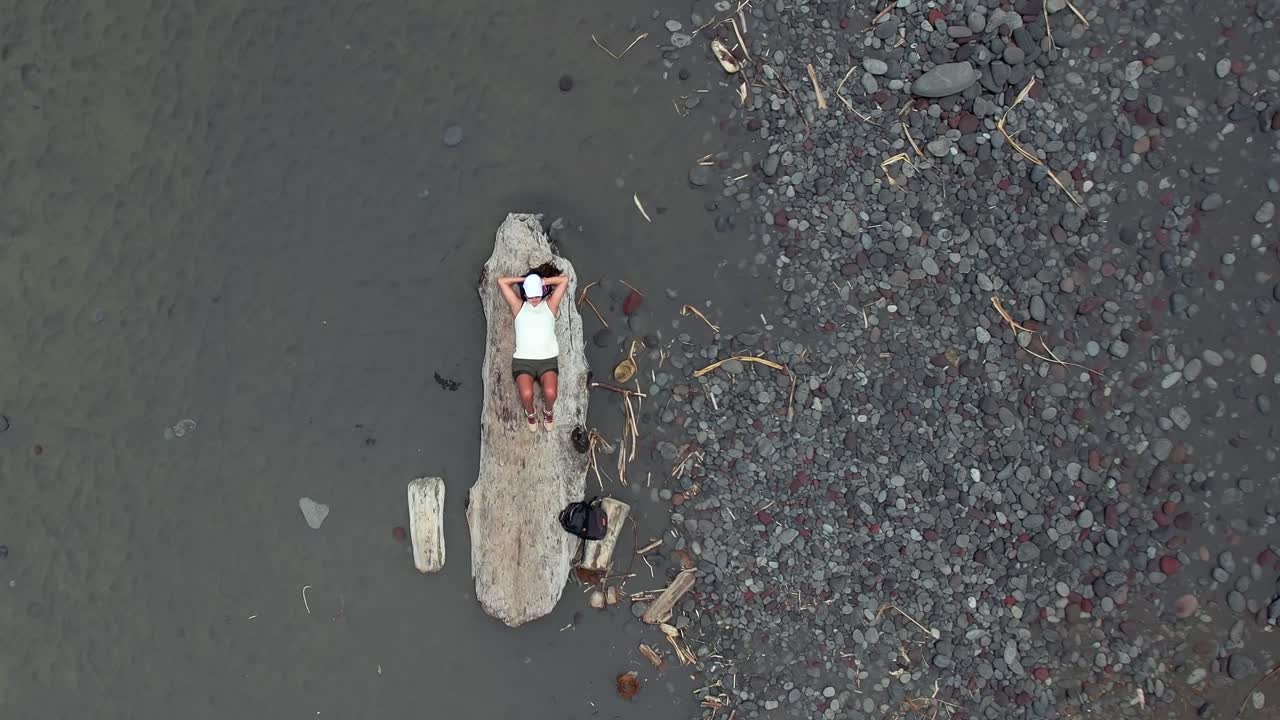 Beautiful young woman relaxing on a plank of drift wood on a black sands beach in Hawaii - ascending straight down aerial view