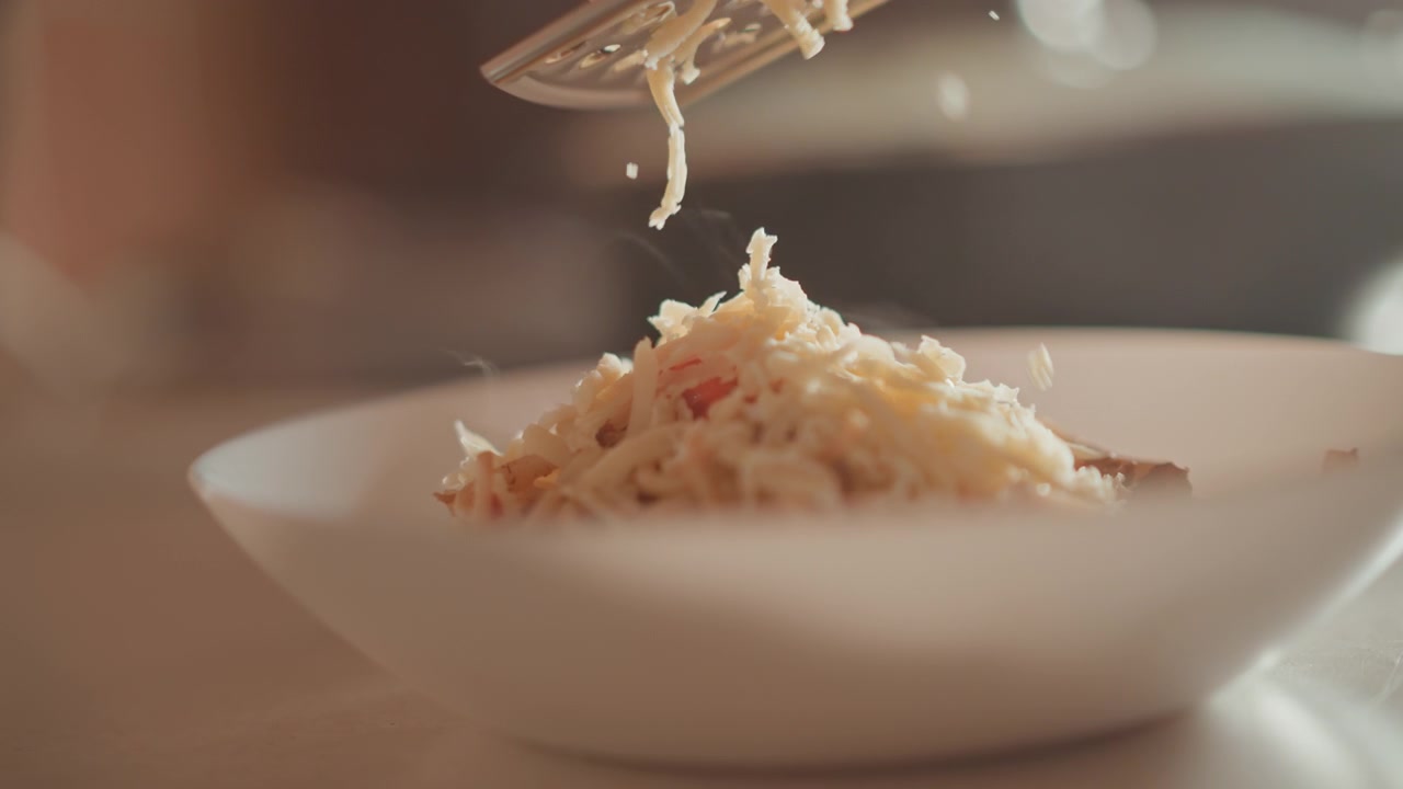 Vista ravvicinata di formaggio grattugiato che si sparge su pasta o risotto impiattati, con filamenti sottili e particelle che si disperdono e si depositano sul cibo cremoso sotto una luce calda e soffusa e una messa a fuoco ridotta.
