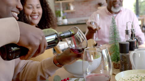 happy african american husband pouring wine for wife and senior father at christmas dinner table