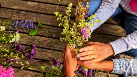 a mother and son create a wildflower bouquet together, their hands arranging fresh spring herbs and flowers on a rustic wooden table, top view