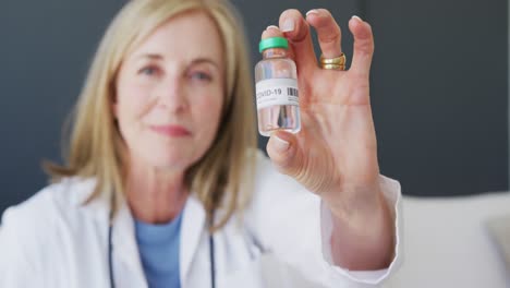 Caucasian-senior-female-doctor-holding-vial-of-coronavirus-vaccine-to-camera-and-smiling