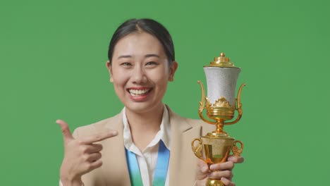 close up of asian business woman in a suit with a gold medal pointing to a gold trophy in her hand being proud winning as the first winner on green screen background in the studio