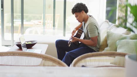 Un-Hombre-Afroamericano-Toca-La-Guitarra-Y-Canta,-Usando-Una-Computadora-Portátil-En-Casa