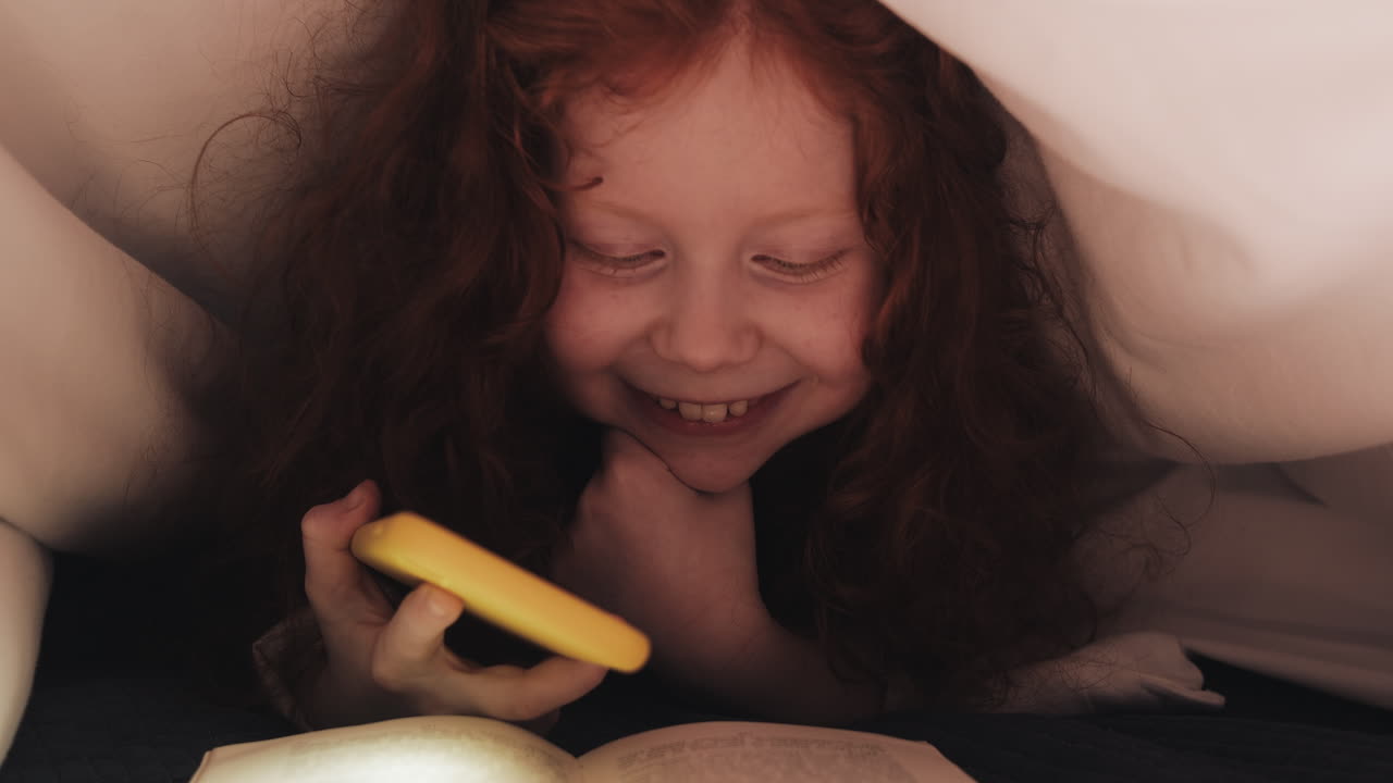 Child Reading Under Blanket with Phone Light