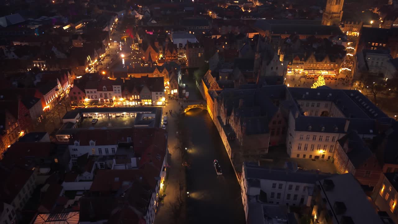 Vista notturna di Bruges decorata per Natale, con canali illuminati e una barca turistica che attraversa il centro città in festa.