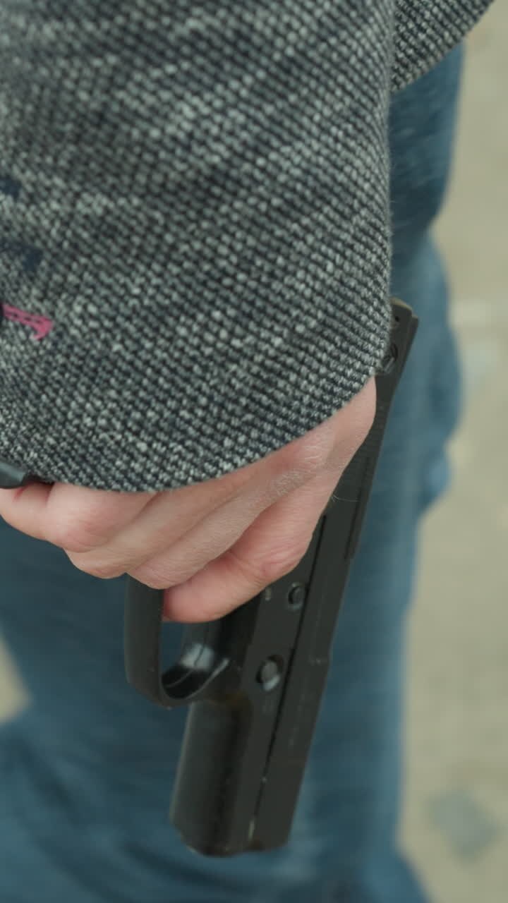 Close view of a person in a gray jacket, black canvas and blue jeans walking on a dirt road, holding a handgun by their side