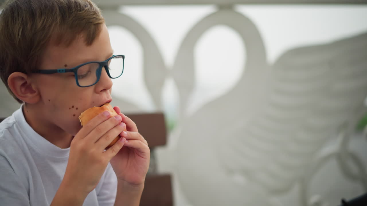 A little boy wearing glasses is seen enjoying his sausage roll while sitting on a bench, he takes a big bite, savoring his snack, with a partial view of another person holding a similar snack