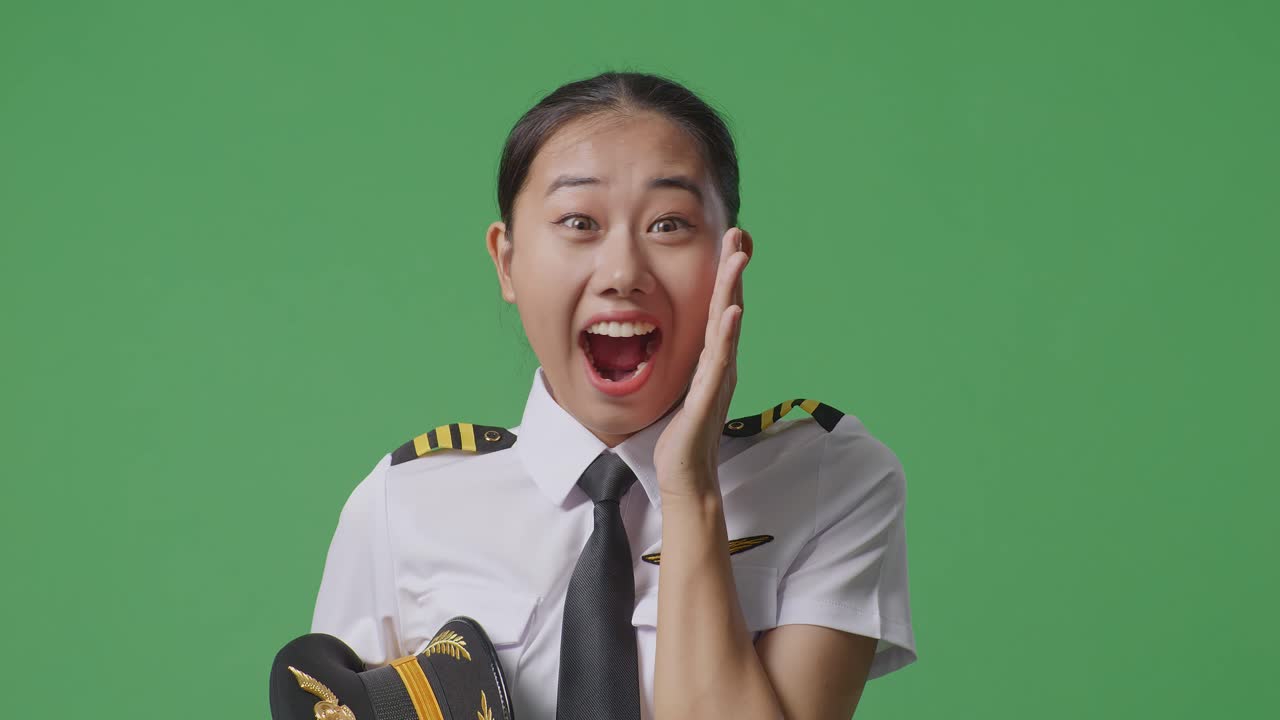 녹색 스크린 배경 스튜디오에 서 있는 아시아 여성 조종사가 미소 짓고 와우라고 말합니다.