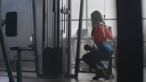 a young student with long hair and a slim body does an exercise on the back muscles on a special training machine. she uses extra load for better results.