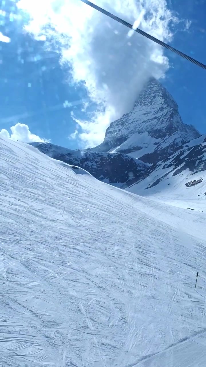 Zermatt'ta alçalan bir teleferik kabini penceresinden düzenlenmiş kayak pistlerinin üzerinden ikonik bulutlarla kaplı Matterhorn zirvesine doğru bakan dikey POV çekimi.