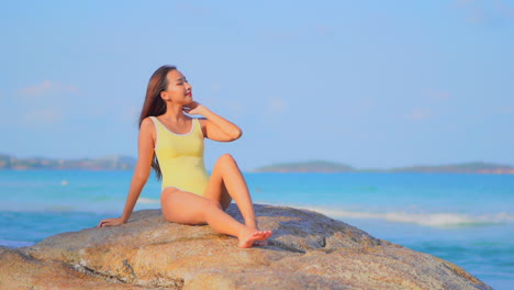 Schöne-Asiatische-Frau-In-Gelbem-Monokini,-Die-Auf-Einem-Großen-Felsen-Am-Meer-Auf-Der-Philippinischen-Insel-Sitzt-Und-Ihr-Haar-Berührt,-Handheld-In-Zeitlupe