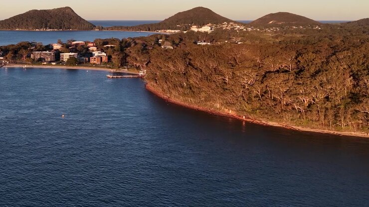 Tomada aérea de drone na hora dourada sobre a costa de Nelson Bay com colinas arborizadas e água calma