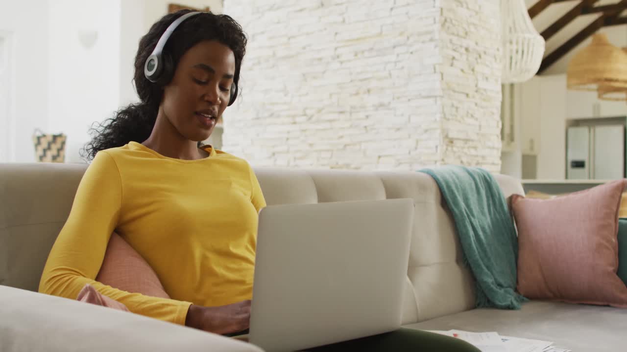 Video of happy african american woman on sofa with headphones using laptop