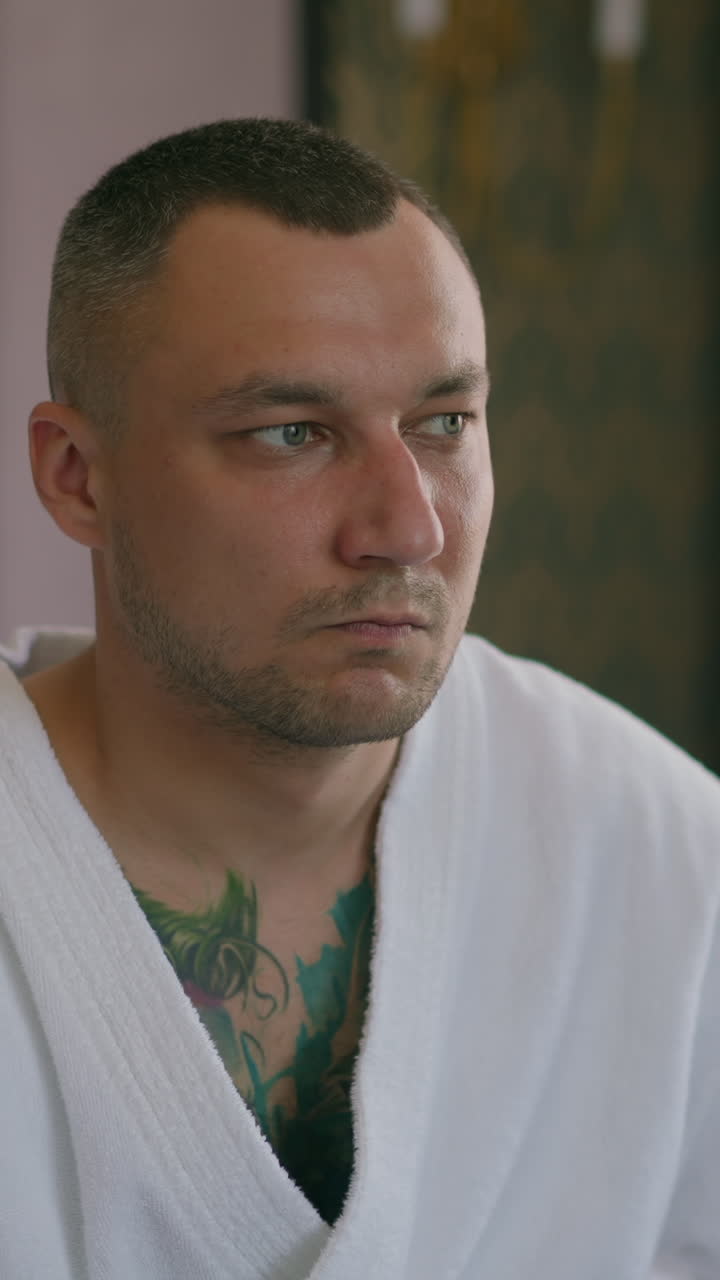 Thoughtful man guest with tattoos on chest in terry bathrobe sits on bed in comfortable hotel room in morning close view slow motion