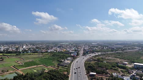 an aerial view of a modern highway flows from green outskirts into urban chennai. busy traffic amidst residential buildings and fields, highlighting regional development and connectivity cloudy sky