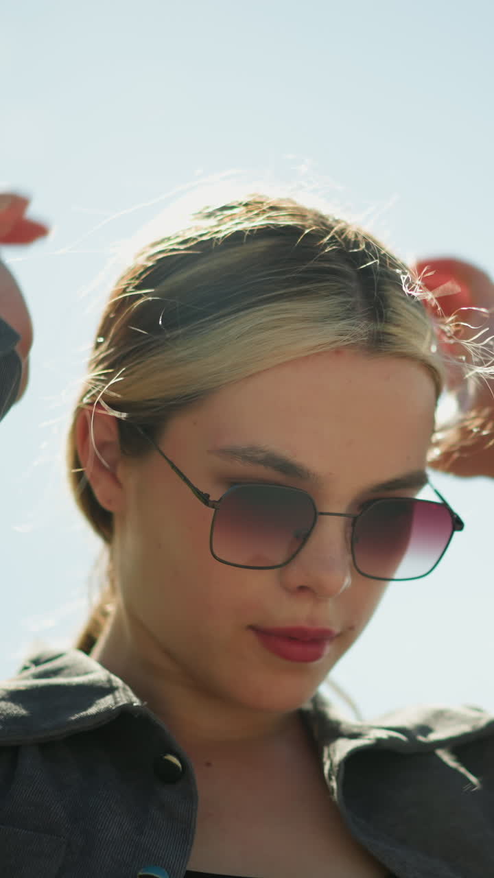 женщина в черном топ-костюме под серой курткой и солнцезащитными очками мягко касается своих волос, когда ветер дует, под ярким, солнечным небом, с волосами, летящими в ветре