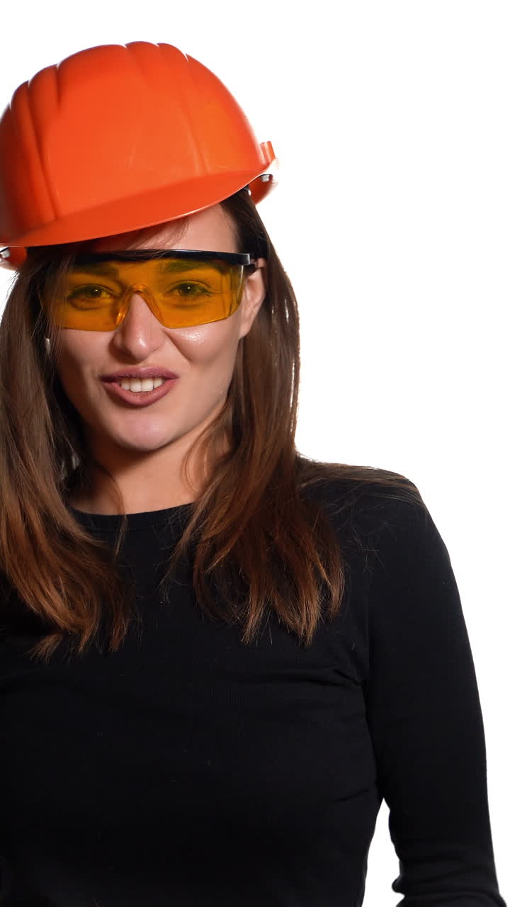 Joyful woman engineer in spectacles is standing in an orange helmet and posing on a white background. Vertical video