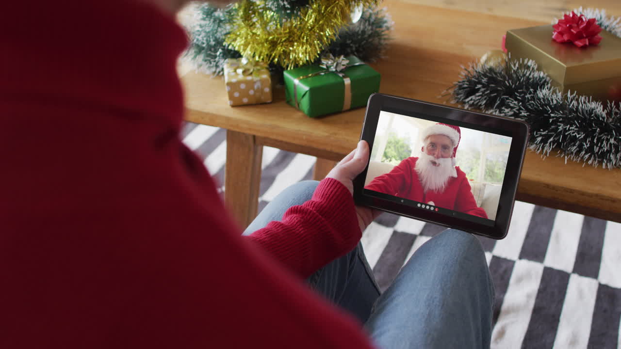 Uomo caucasico che saluta con la mano e usa un tablet per una videochiamata di Natale con Babbo Natale sorridente sullo schermo.