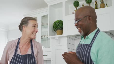 Smiling-senior-diverse-couple-wearing-blue-aprons-and-cooking-in-kitchen