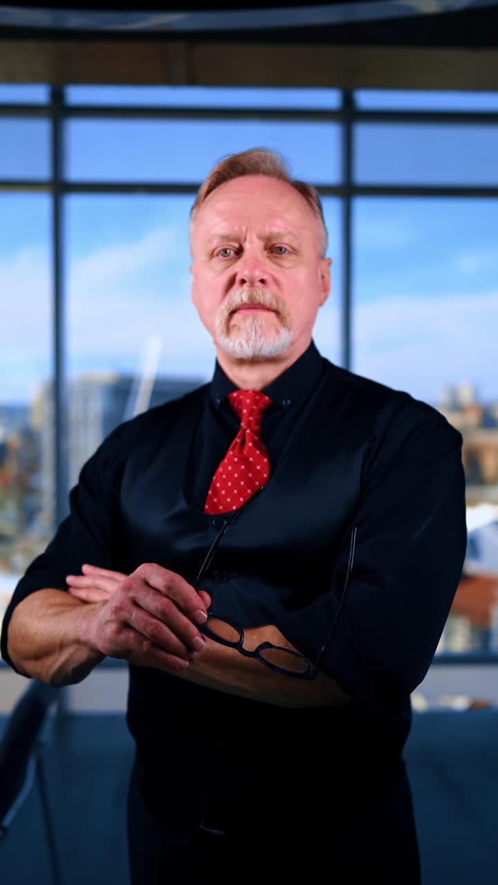 Bearded Caucasian male in dark shirt, vest and red tie stands his arm folded on his chest. Man puts on glasses and stands still in front of camera. Vertical video.