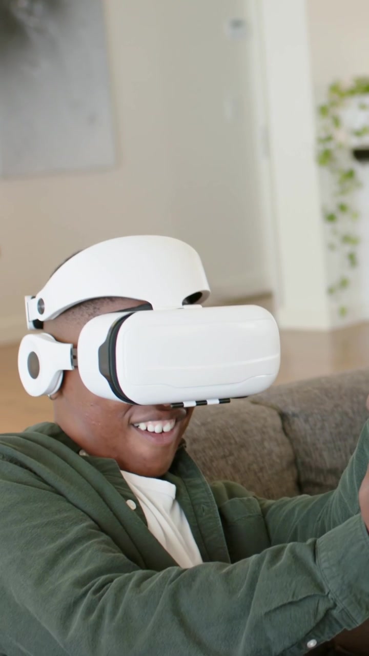 Video verticale: Adolescente sorridente in verde che avvia un visore VR, alzando il controller sul divano e giocando a un videogioco.