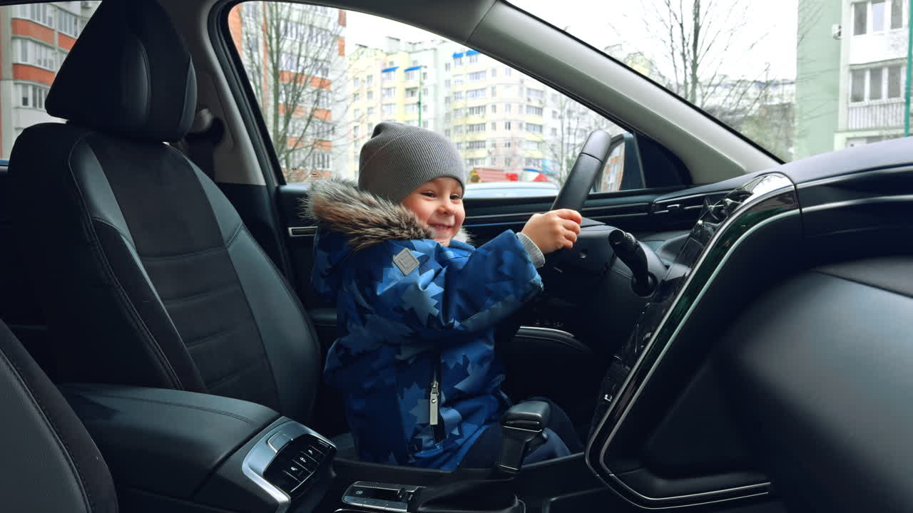 Un allegro bambino caucasico con giacca calda e berretto grigio è in macchina sul sedile del conducente. Un bambino felice e sorridente si tiene al volante. Primo piano.