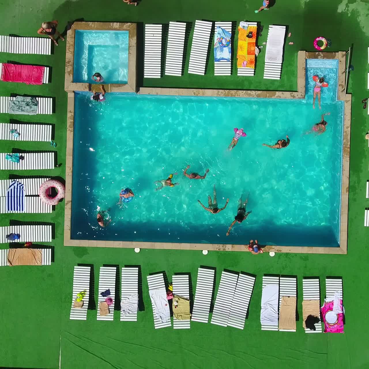 Happy people swimming in the blue pool, doing aerobics around and sunbathing on the chaise-longues. Drone footage going up over the spa resort territory