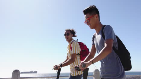happy diverse gay male couple using scooters at promenade by the sea, slow motion