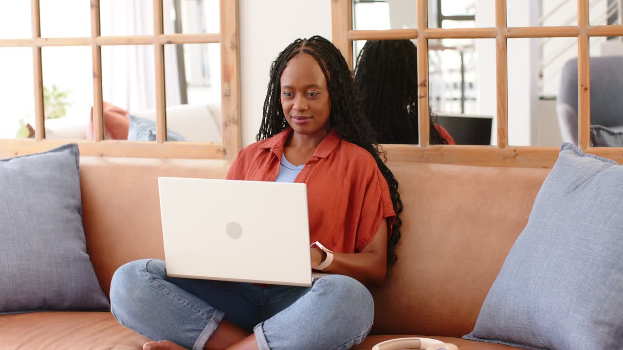 Adult African woman responding to screen content, typing on white laptop while sitting on tan sofa