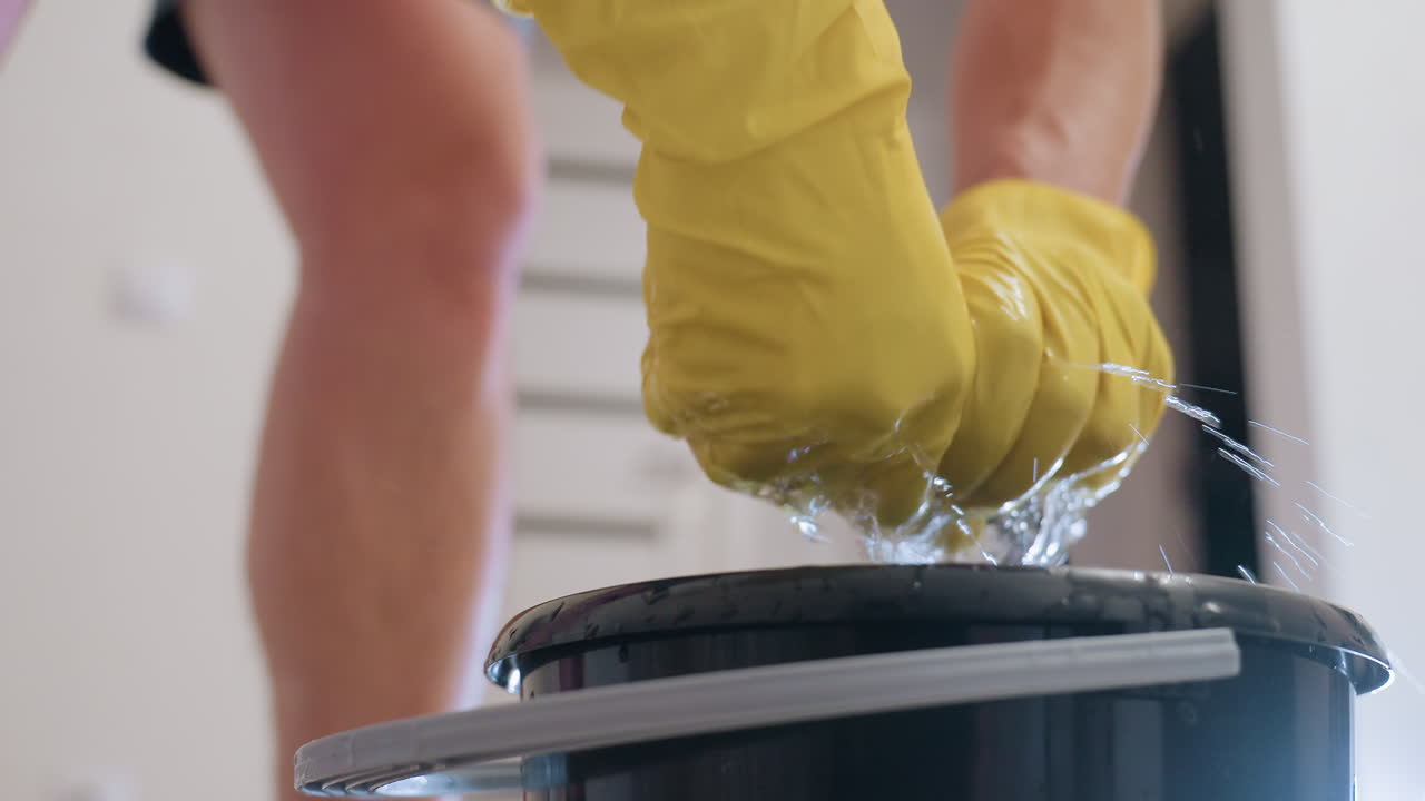 Close up of cleaner wearing yellow gloves lifting black bucket on wooden floor during household chores, showing daily cleaning task with sanitation effort, manual work and hygiene detail
