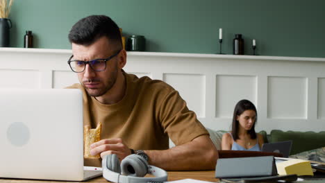 close up view of a student with headphones, using laptop and eating a sandwich sitting at table