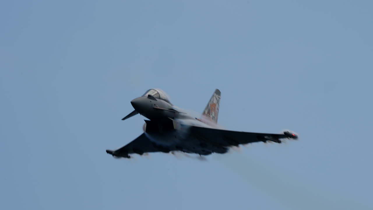 Military jet aircraft flying fast and low with visible vapor trails around the wings against clear blue sky