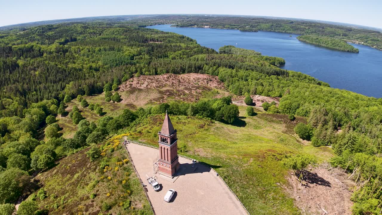 Drone footage shows a tall brick tower on a forested peninsula by a lake in Denmark. The camera circles above the historic structure, revealing surrounding green trees and serene water