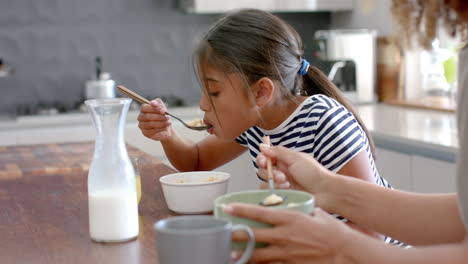 Biracial-mother-and-daughter-eating-breakfast-cereal-in-sunny-kitchen,-slow-motion