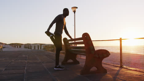 hombre afroamericano enfocado estirándose en un banco, haciendo ejercicio al aire libre en la orilla del mar al atardecer